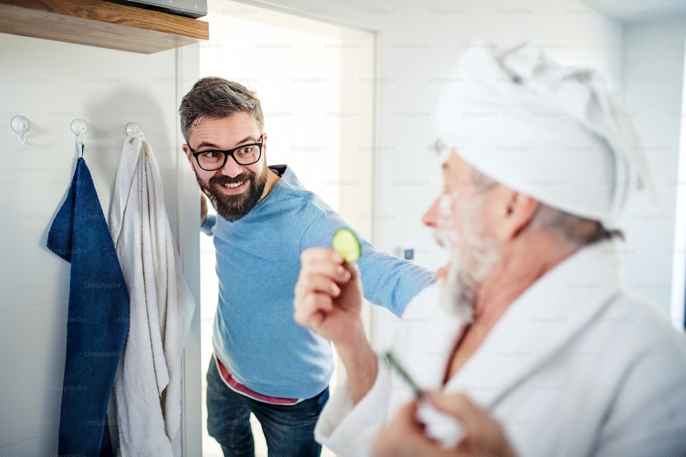 A cheerful adult hipster son and senior father in bathroom indoors at home, having fun.