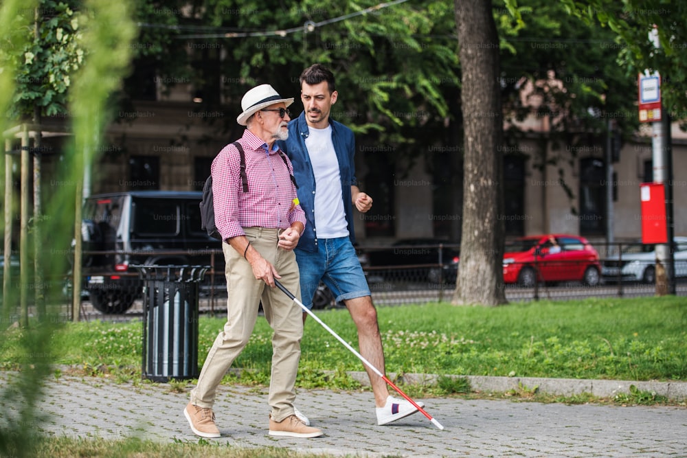 Un hombre joven y un anciano ciego con bastón blanco caminando por la acera en la ciudad, hablando.