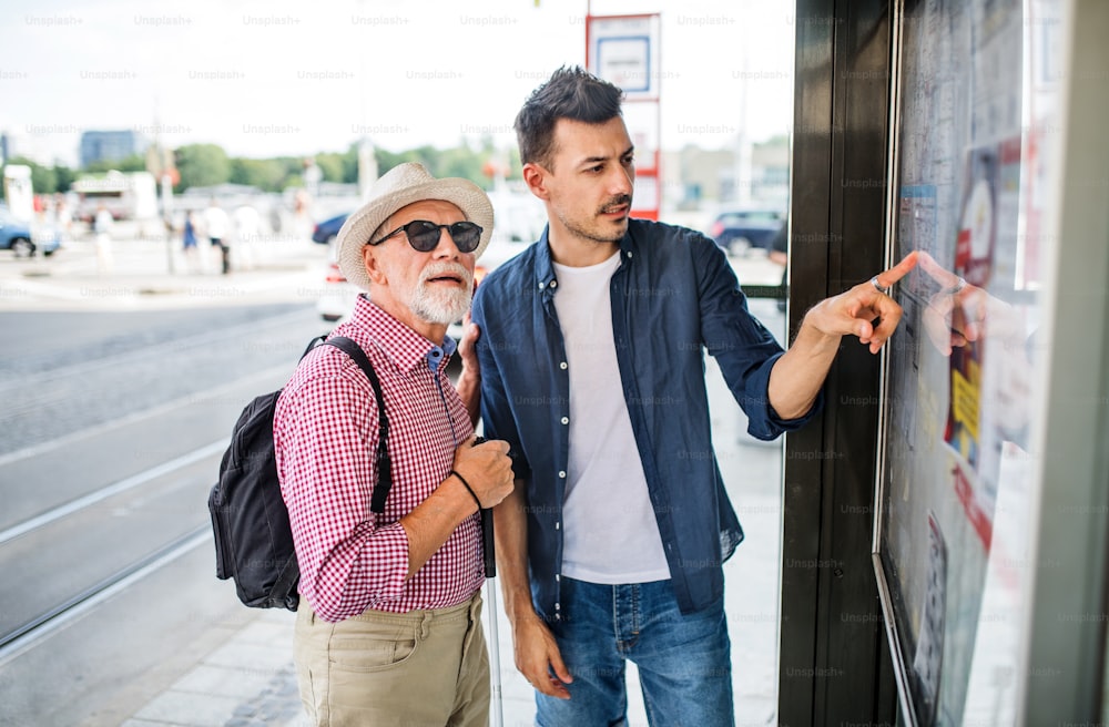 A young man and blind senior with white cane at bus stop in city, reading timetable.