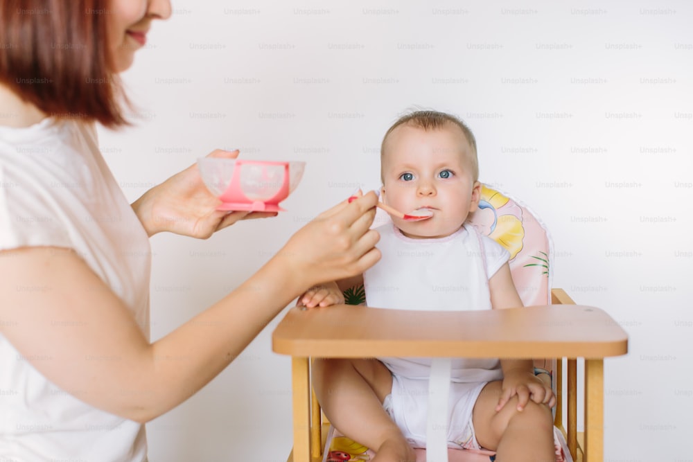 Mother Feeding Baby Sitting In High Chair At Mealtime