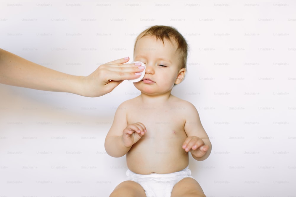 Une mère nettoie le visage d’une fille nouveau-née avec un disque ouaté isolé sur fond blanc. Hygiène, concept de soins de santé.