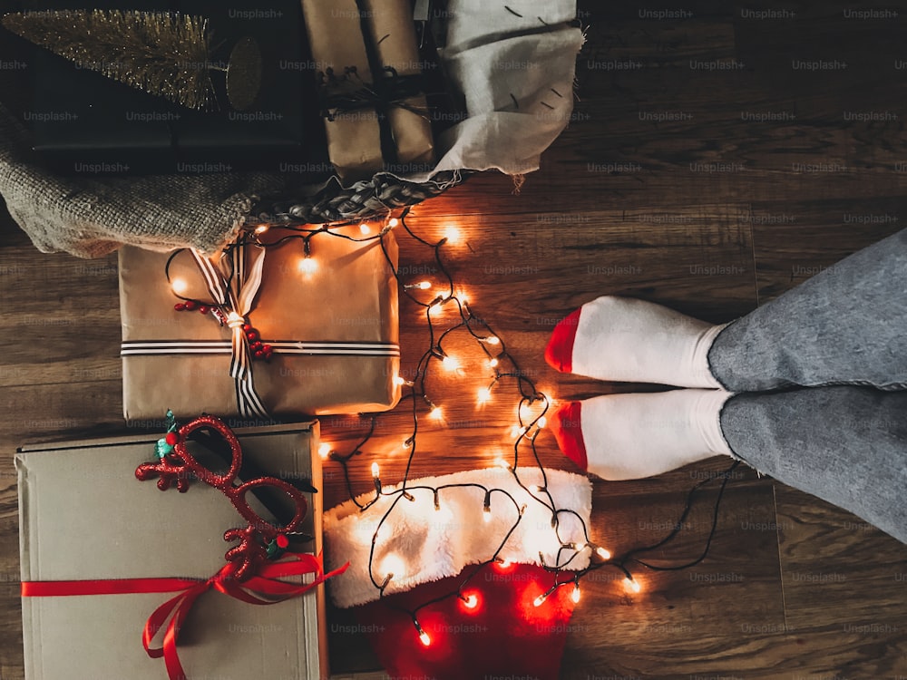 Coffrets cadeaux de Noël, lumières festives, bonnet de père Noël et jambes de fille dans des chaussettes douillettes sur un plancher en bois la veille de Noël. Joyeuses Fêtes. Vue de dessus. Photo du téléphone