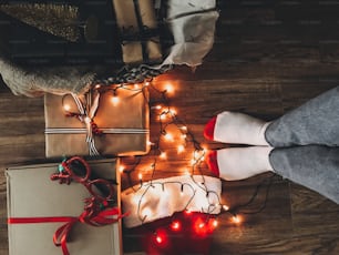 Cajas de regalo de Navidad, luces festivas, gorro de Papá Noel y piernas de niña en calcetines acogedores sobre piso de madera en Nochebuena. Felices Fiestas. Vista superior. Foto del teléfono