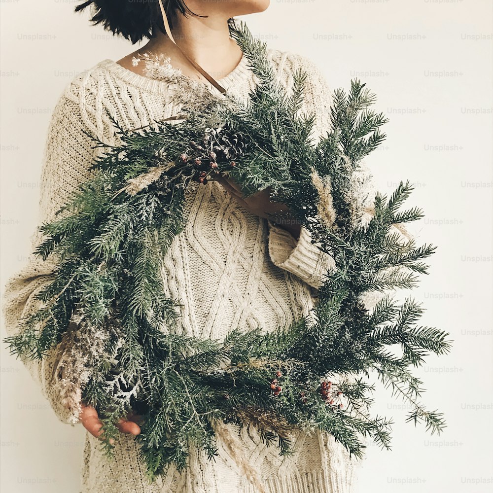 Stylish rustic wreath in girl hands. Hipster girl in knitted sweater holding rural modern wreath on white wall background. Festive decoration. Phone photo