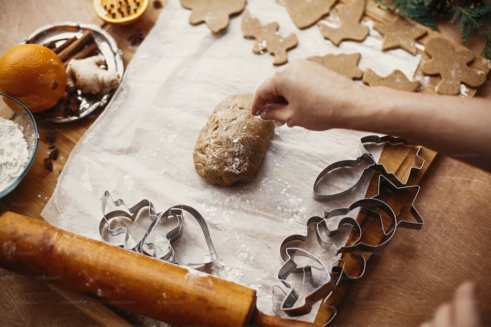 Fare biscotti di pan di zenzero di Natale. Mani che impastano pasta cruda su sfondo di mattarello, taglierine metalliche, anice, zenzero, cannella, pigne, rami di abete su tavolo rustico.