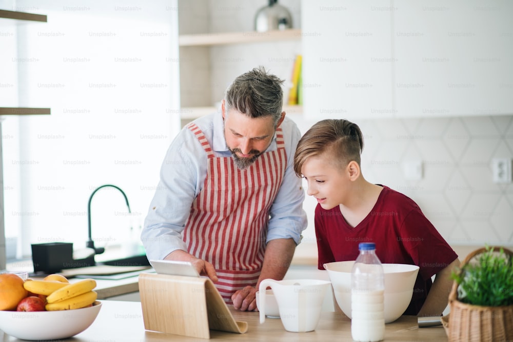 Um pai maduro e um filho pequeno com tablet dentro de casa na cozinha, fazendo panquecas.