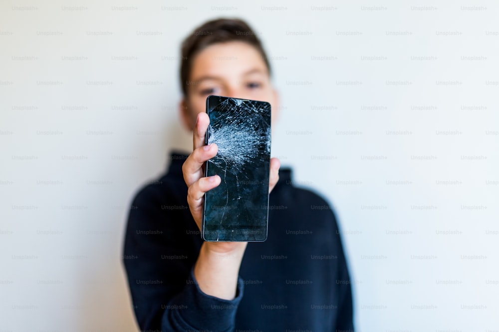 Teléfono inteligente con pantalla de vidrio roto en la mano de un niño molesto, fondo blanco. Niño preocupado sosteniendo un teléfono inteligente roto en casa