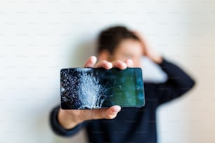 Teléfono inteligente con pantalla de vidrio roto en la mano de un niño molesto, fondo blanco. Niño preocupado sosteniendo un teléfono inteligente roto en casa