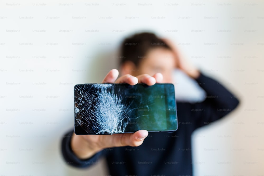 動揺した少年の手にある壊れたガラス画面のスマートフォン、白い背景。家で壊れたスマートフォンを抱きしめている心配な子供