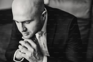 Stylish groom in black suit getting ready in the morning, preparing for wedding ceremony in room. Confident bald man sitting in chair. Businessman in suit. Black white photo