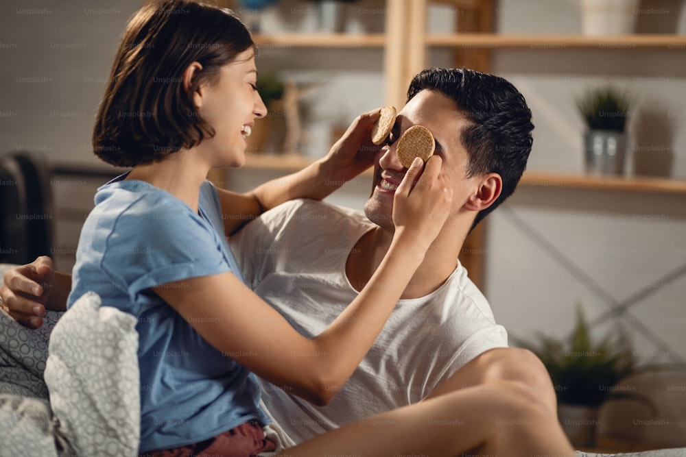 Happy couple having fun with food. Young woman is holding cookies and covering boyfriend eyes.