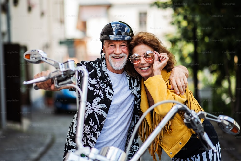 A front view of cheerful senior couple travellers with motorbike in town.