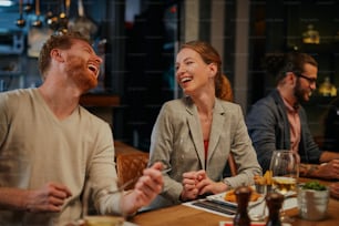 Charming smiling caucasian blonde sitting in restaurant, eating dinner and chatting with friend. In background are friends.