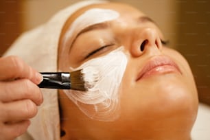Close-up de esteticista aplicando máscara branca no rosto da mulher durante o tratamento de beleza no spa.