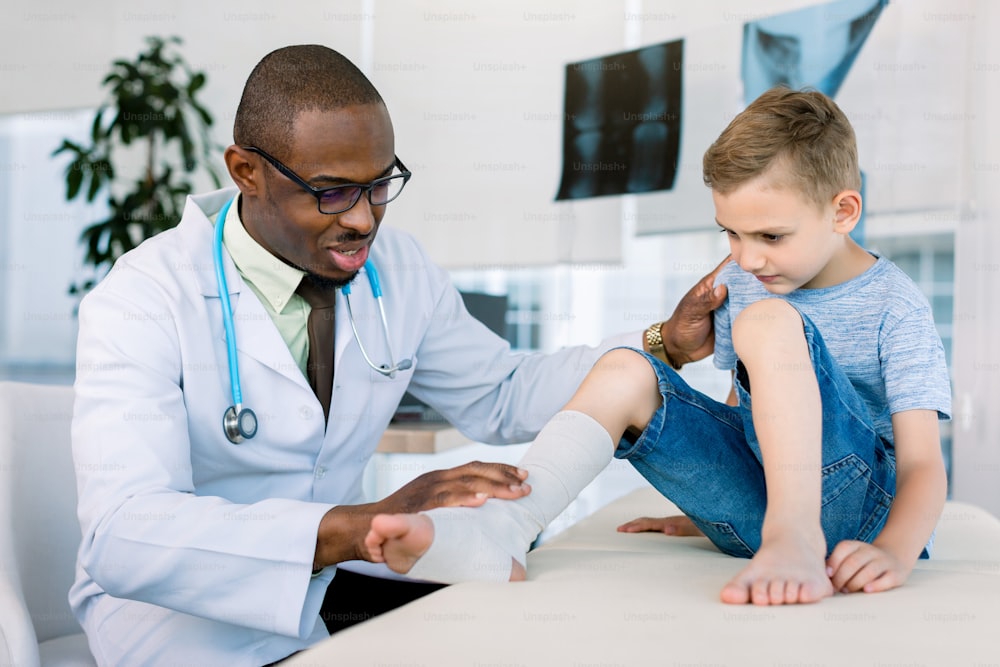 Leg injured boy visiting young African doctor traumatologist in his medical clinic hospital. Boy is sitting on the couch. Doctor in white coat examines the leg.