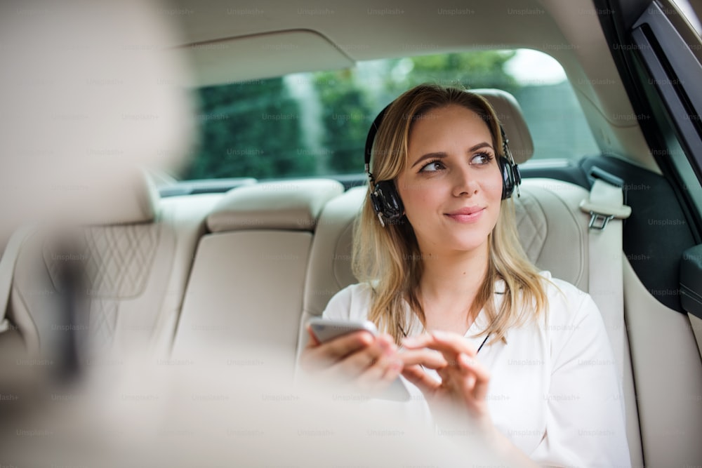 Business woman with smartphone and headphones sitting on back seats in taxi car, listening to music.