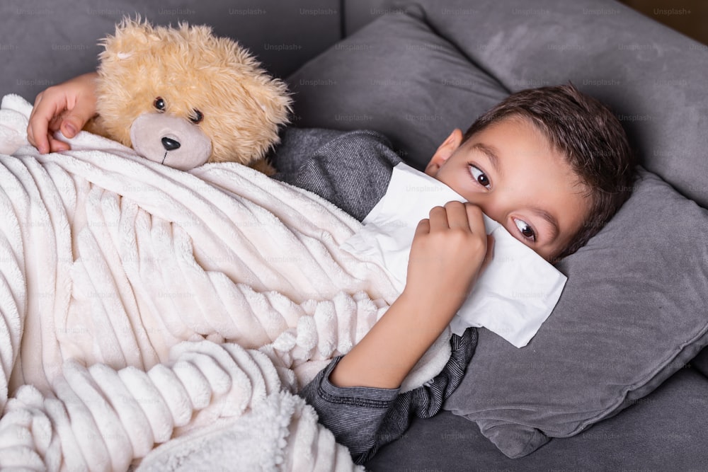 Resfriado e gripe. Retrato de menino doente pego frio, sentindo-se doente e espirrando em papel limpo. Closeup de criança insalubre jovem coberta de cobertor limpando o nariz. Conceito de Saúde.