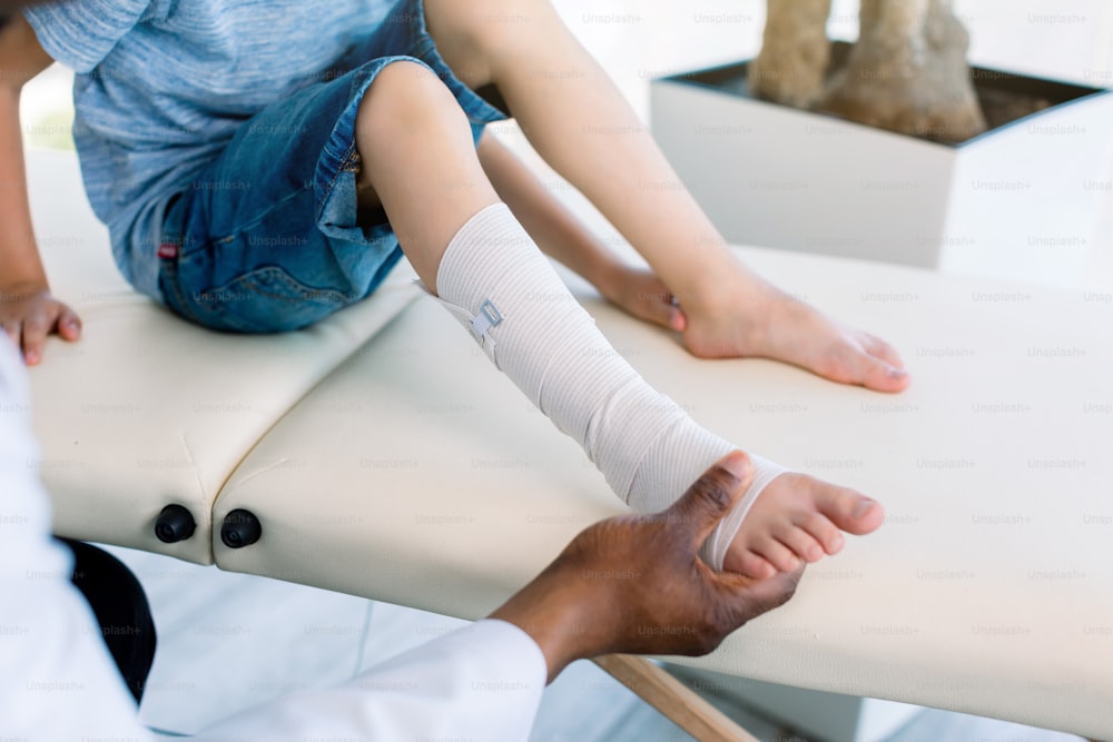 cropped shot of african american doctor putting on bandage on patients injured leg. Doctor first aid, child patient's leg sprain with elastic bandage