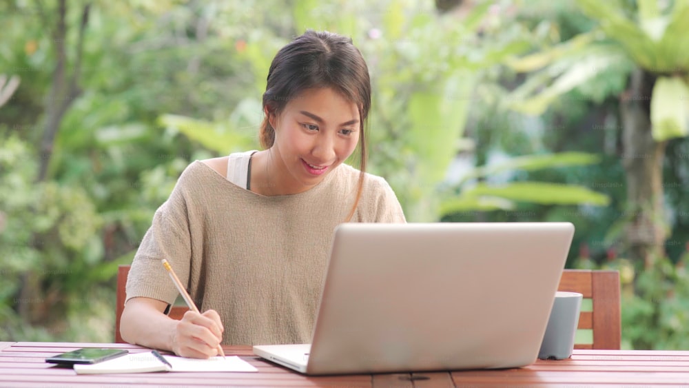 Freelance Asian woman working at home, business female working on laptop sitting on table in the garden in morning. Lifestyle women working at home concept.