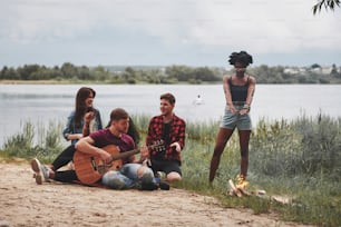 Uomo in camicia rossa che canta una canzone. Un gruppo di persone fa un picnic sulla spiaggia. Gli amici si divertono nel fine settimana.