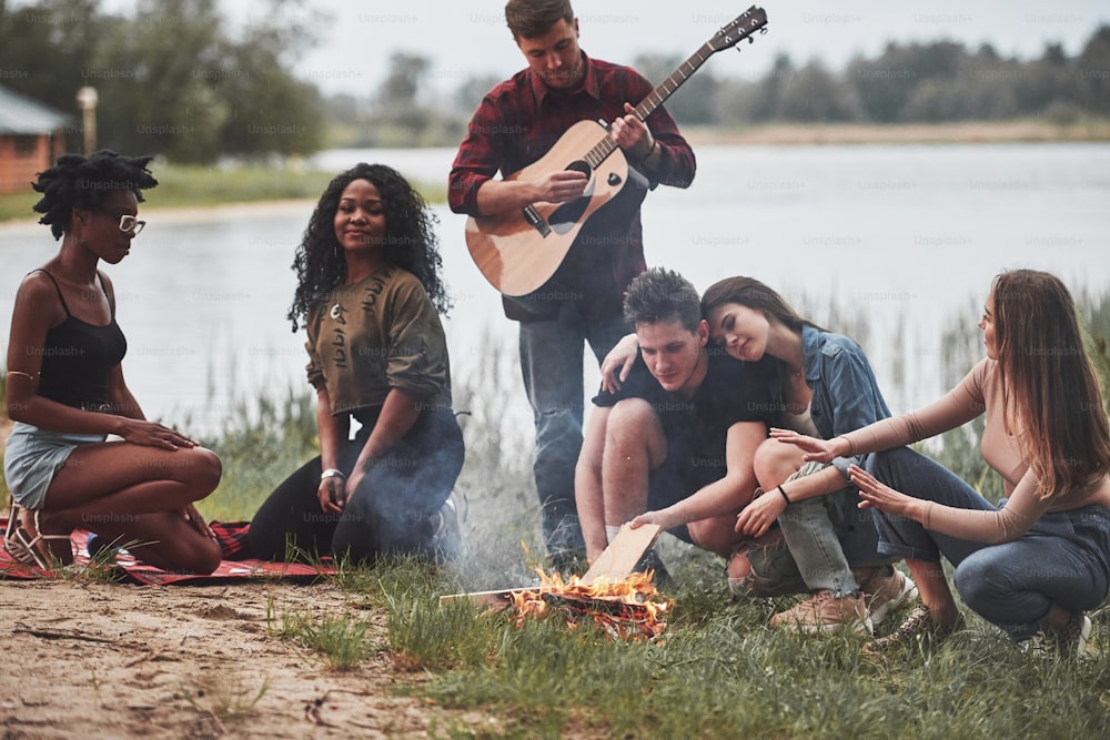 Standing with guitar. Group of people have picnic on the beach. Friends have fun at weekend time.