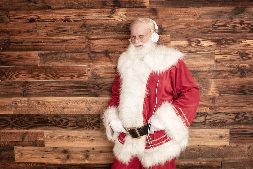 Sonriente verdadero Santa Claus escuchando música con auriculares, posando en la pared de madera. Feliz Navidad y Feliz Año Nuevo concepto. Emociones y expresión.