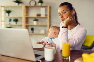 Young displeased mother surfing the net on a computer while being with her baby a t home.