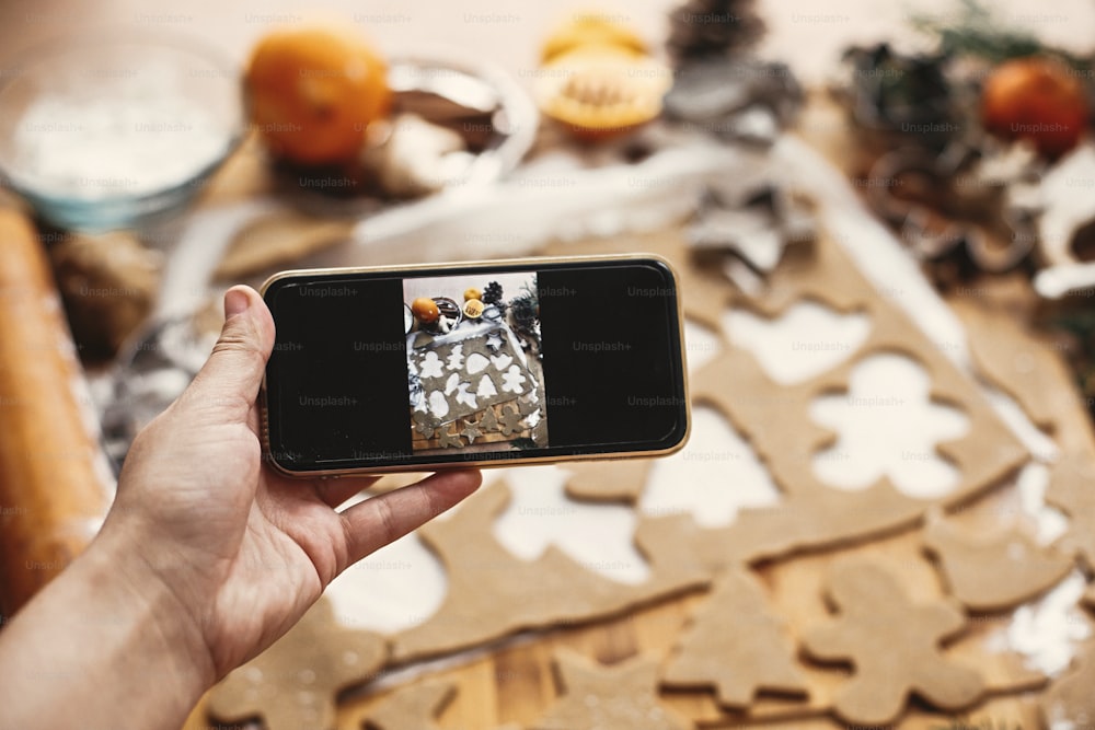 Mano che tiene il telefono e scatta foto del processo di preparazione dei biscotti di pan di zenzero di Natale. Scattare foto sullo smartphone di biscotti di pan di zenzero crudi sul tavolo rustico.