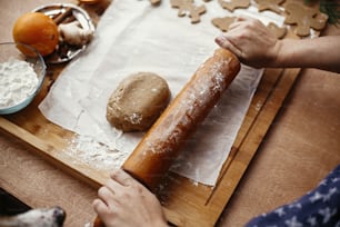 素朴なテーブルの上の金属カッター、アニス、生姜、シナモン、松ぼっくり、モミの枝の背景に木製の麺棒で生生地を転がす手。クリスマスのジンジャーブレッドクッキーを作ります。