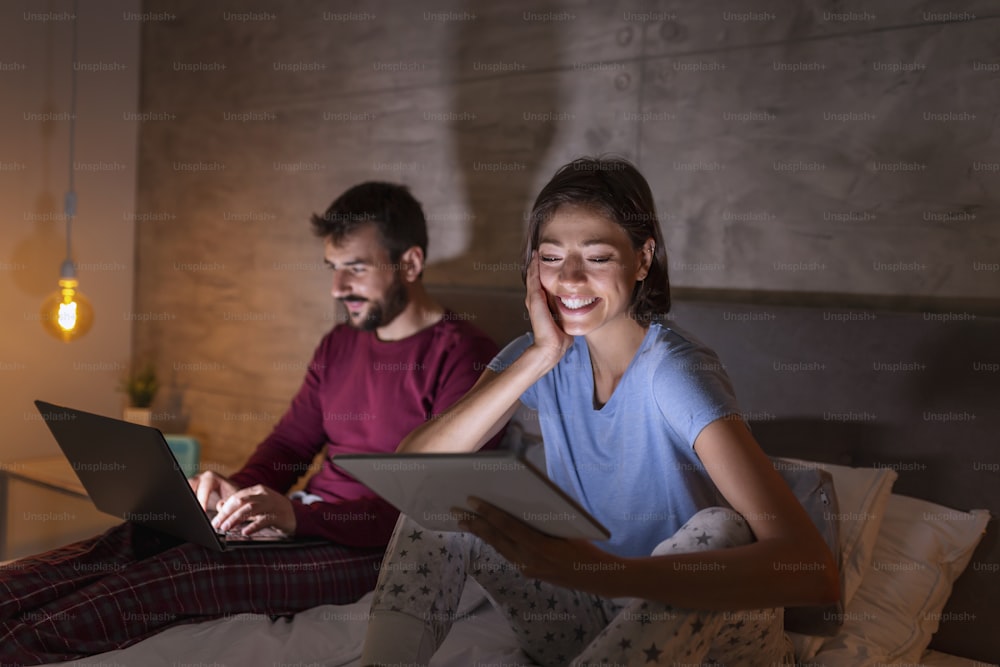 Beautiful young couple sitting side by side in bed at night, reading and watching TV shows on laptop and tablet computers