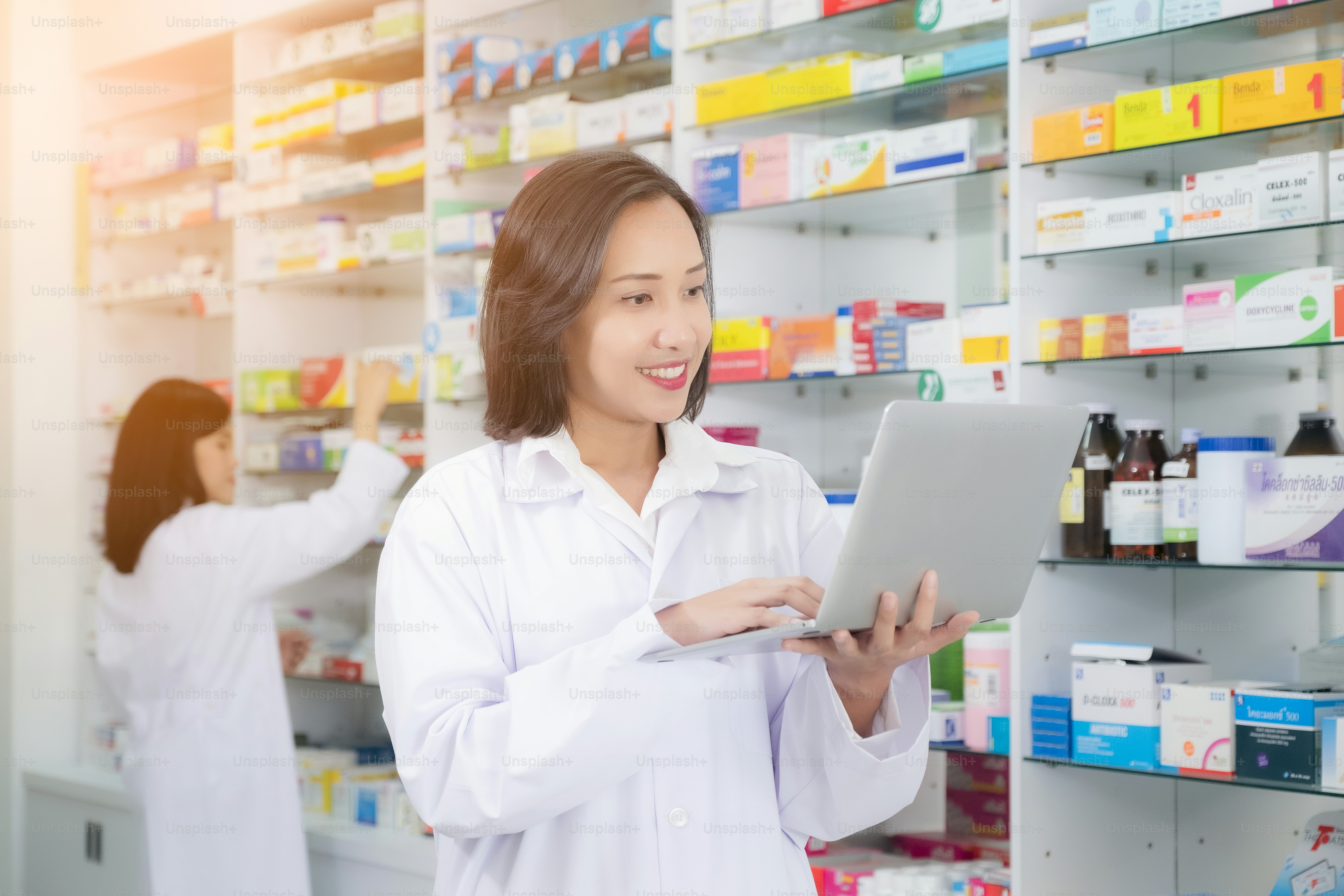 Asiatische Apothekerin weiblich mit Laptop-Computer und Checkliste Inventar oder Lesen von Informationen Medikamente in Apotheke oder Drogerie. Medizin, Pharmazie, Gesundheitswesen und Menschenkonzept