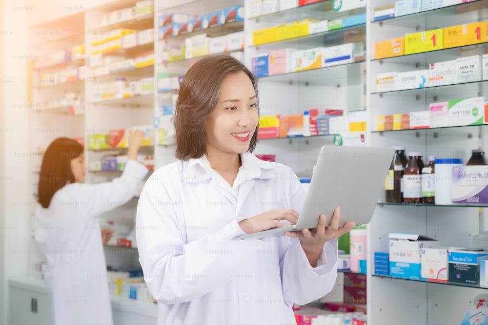Asian pharmacist female using laptop computer and checklist inventory or reading information medication in pharmacy or drugstore. medicine, pharmaceutics, health care and people concept