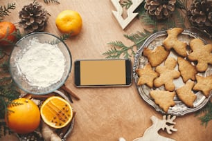빈 화면과 아니스, 계피, 솔방울, 소박한 테이블에 삼나무 가지, 평평한 누워 있는 축제 진저브레드 쿠키가 있는 전화. 텍스트를 위한 공간입니다. 즐거운 성탄절. 휴일 카드 모형