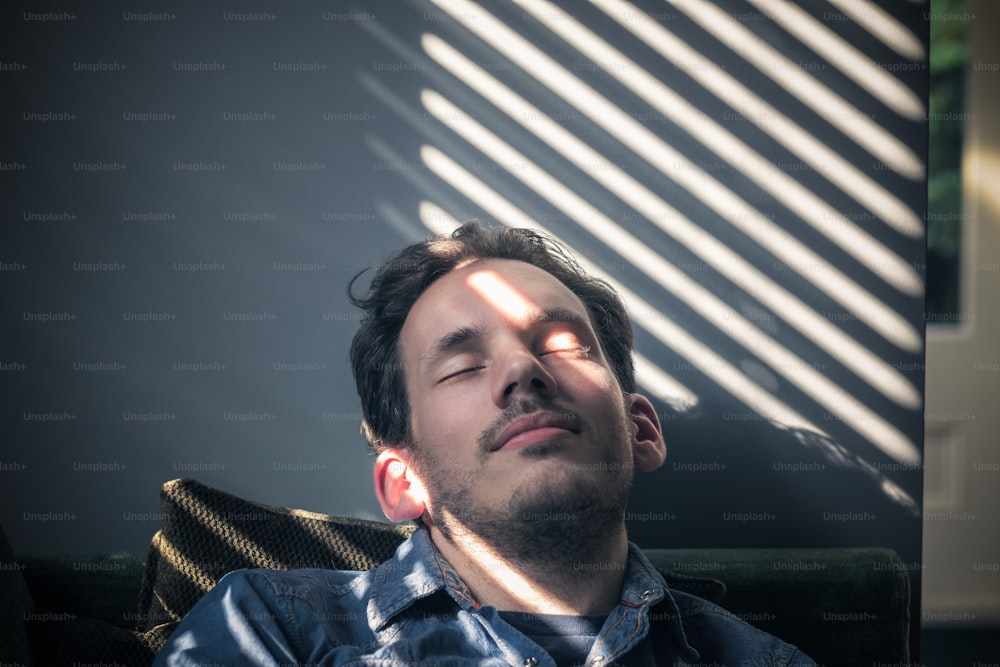 Portrait d’un homme caucasien somnolant sur le canapé, sur le visage tombe l’ombre des stores