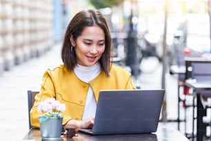 Young asian business woman in dress sitting at table in cafe and writing in notebook. On table is laptop, smartphone and cup of coffee. Freelancer working in coffee shop. Student learning online.
