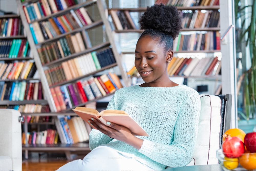 Jeune belle fille afro-américaine lisant un livre sur le canapé avec les étagères de la bibliothèque à l’arrière. Belle femme sur un canapé blanc lisant un livre