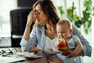 Young working mother having a headache and feeling tired while working at home and taking care of her baby son. Focus is on crying baby.