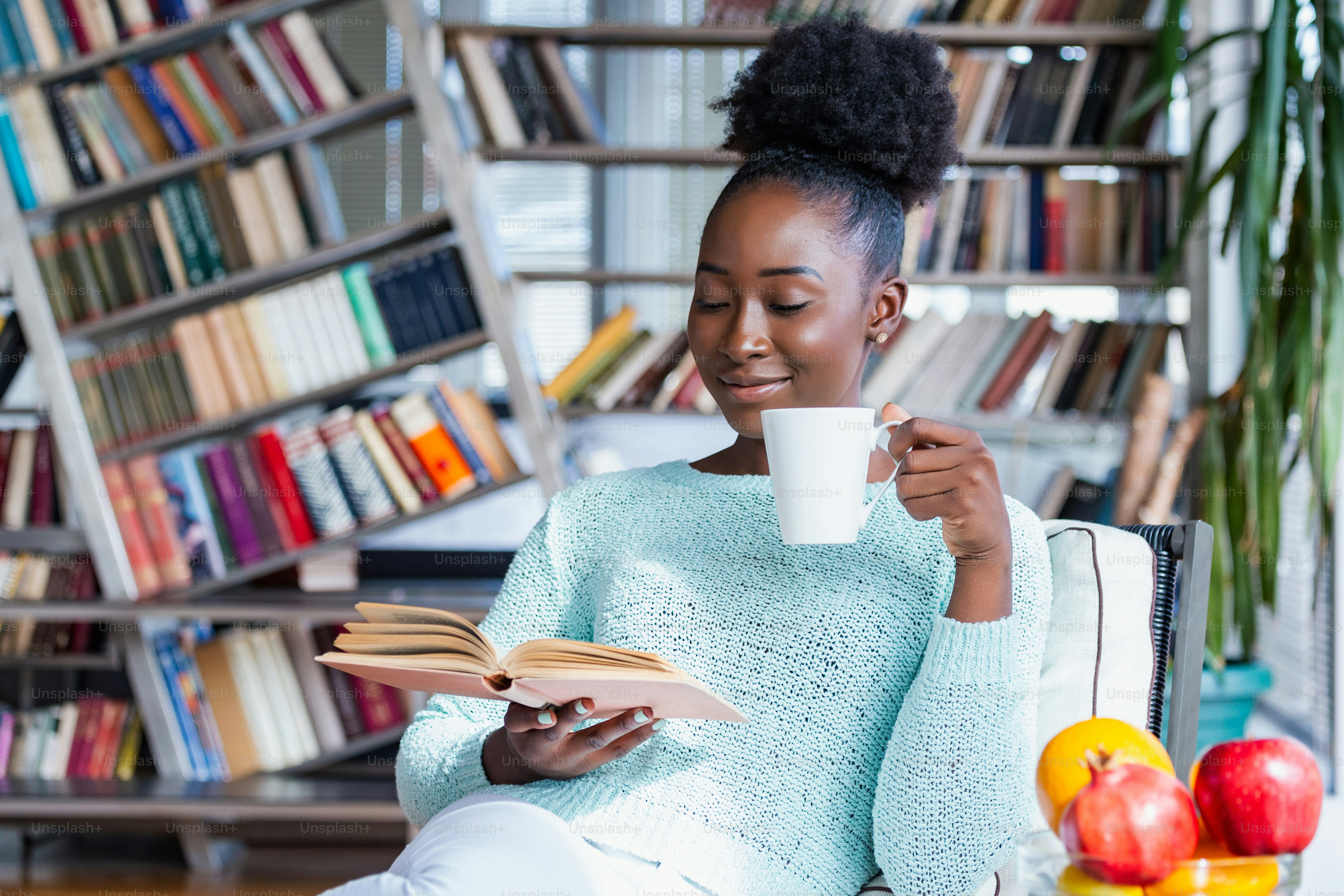 A woman in a light blue sweater reading a book while holding a mug