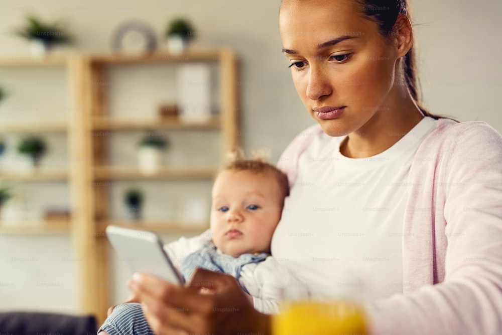 Pensive mother using cell phone and typing text message while being with her baby at home.