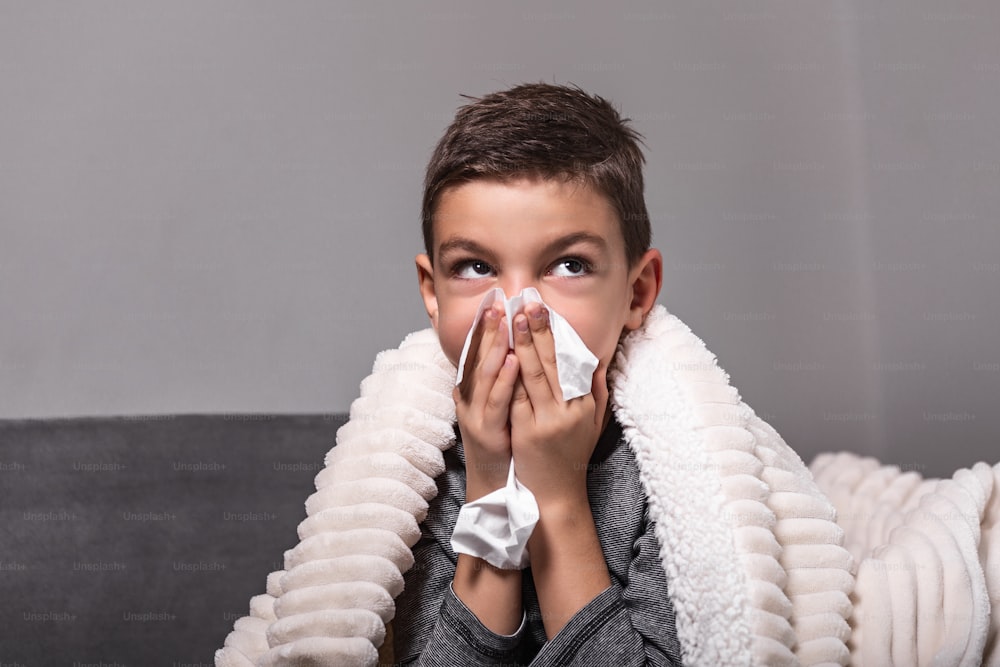 Resfriado e gripe. Retrato de menino doente pego frio, sentindo-se doente e espirrando em papel limpo. Closeup de criança insalubre jovem coberta de cobertor limpando o nariz. Conceito de Saúde.