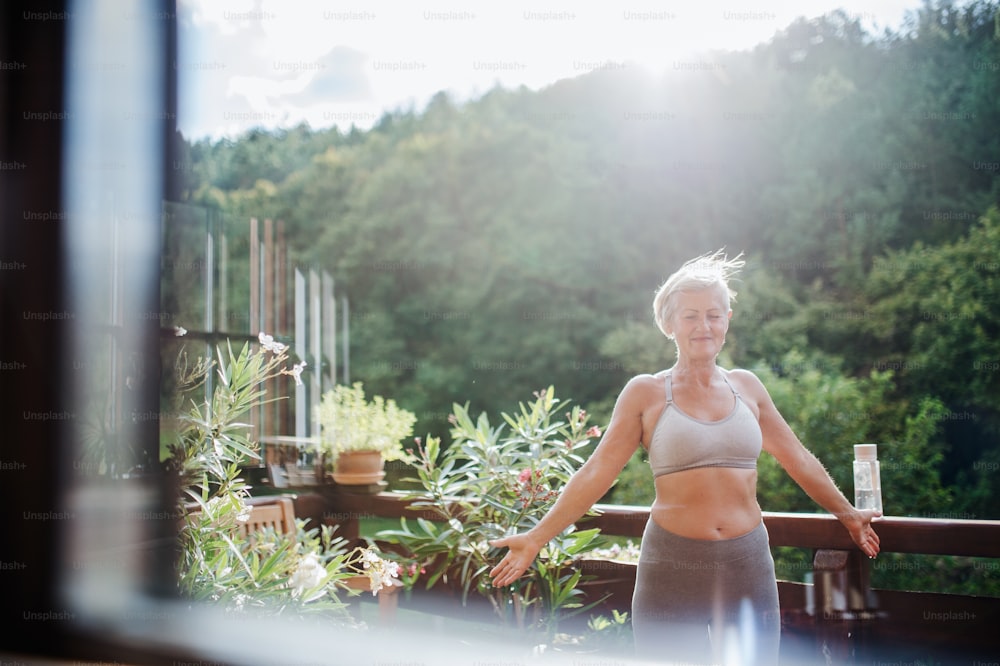 A senior woman with sports bra outdoors on a terrace in summer, doing exercise. Shot through glass.