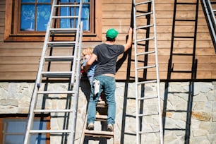 夏の屋外で木造住宅を塗装する父親と幼児の男の子の後ろ姿。