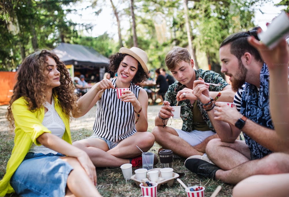 Grupo de jovens amigos alegres no festival de verão, sentados no chão e comendo.