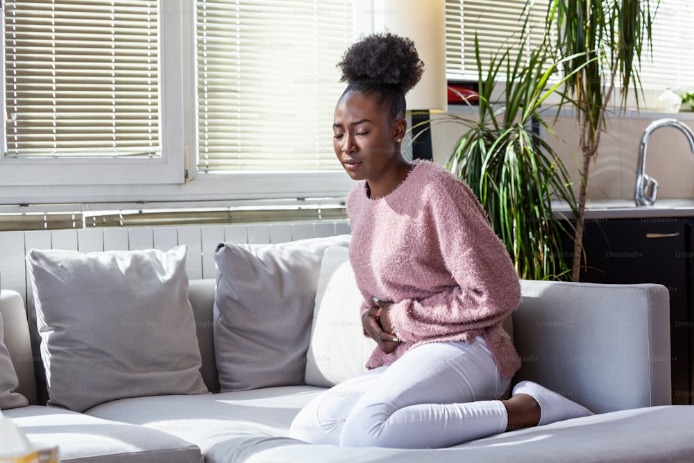 Young black woman suffering from stomachache on sofa at home. Woman sitting on bed and having stomach ache. Young woman suffering from abdominal pain while sitting on sofa at home