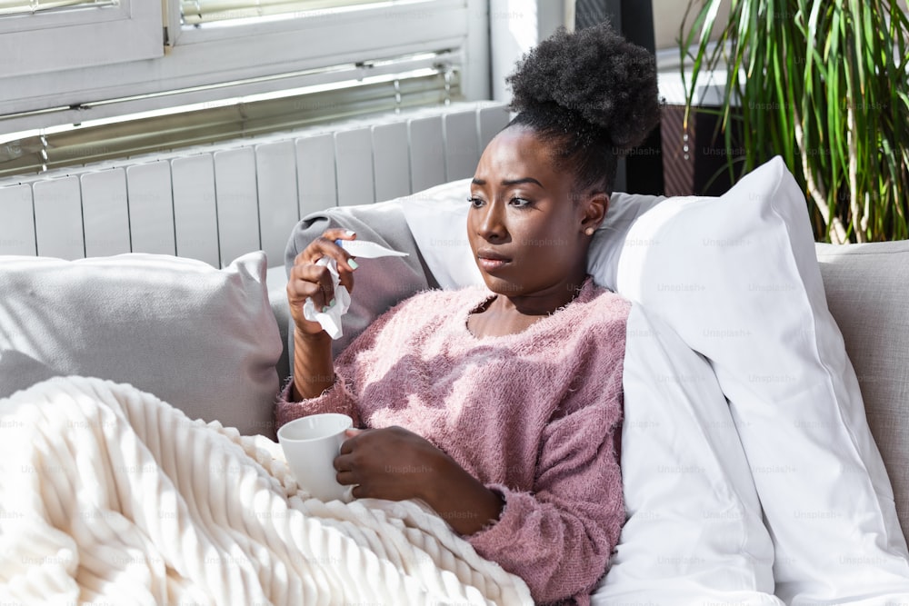 Enfermedad, concepto de problema de virus estacional. Mujer afroamericana enferma que tiene gripe acostada en el sofá mirando la temperatura en el termómetro. Mujer enferma acostada en cama con fiebre alta. Resfriado, gripe y migraña.