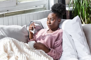 Sickness, seasonal virus problem concept. African American Woman being sick having flu lying on sofa looking at temperature on thermometer. Sick woman lying in bed with high fever. Cold flu and migraine.