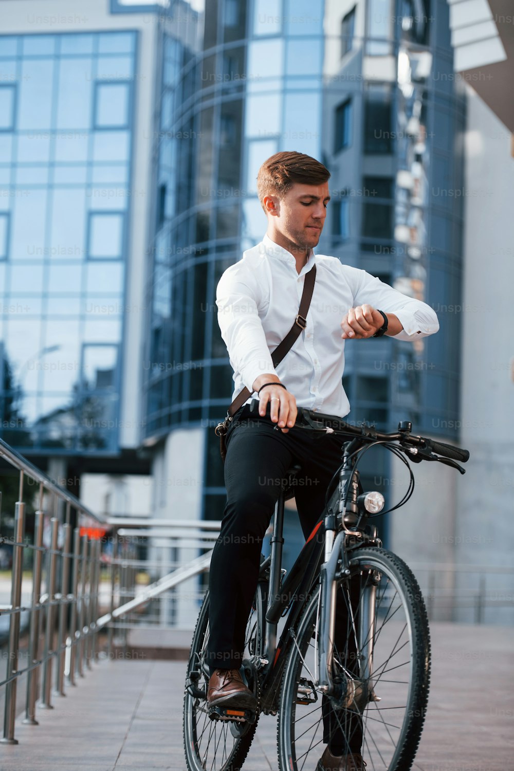 Looks at watch. Businessman in formal clothes with black bicycle is in the city.