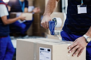 Close-up of delivery man closing carboard box with a tape while preparing packages for shipment.