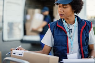 Afroamerikanischer Arbeiter, der den Lieferplan organisiert und vor der Lieferung Kästchen ankreuzt. Ihre Kollegin verlädt im Hintergrund Pakete in einen Transporter.