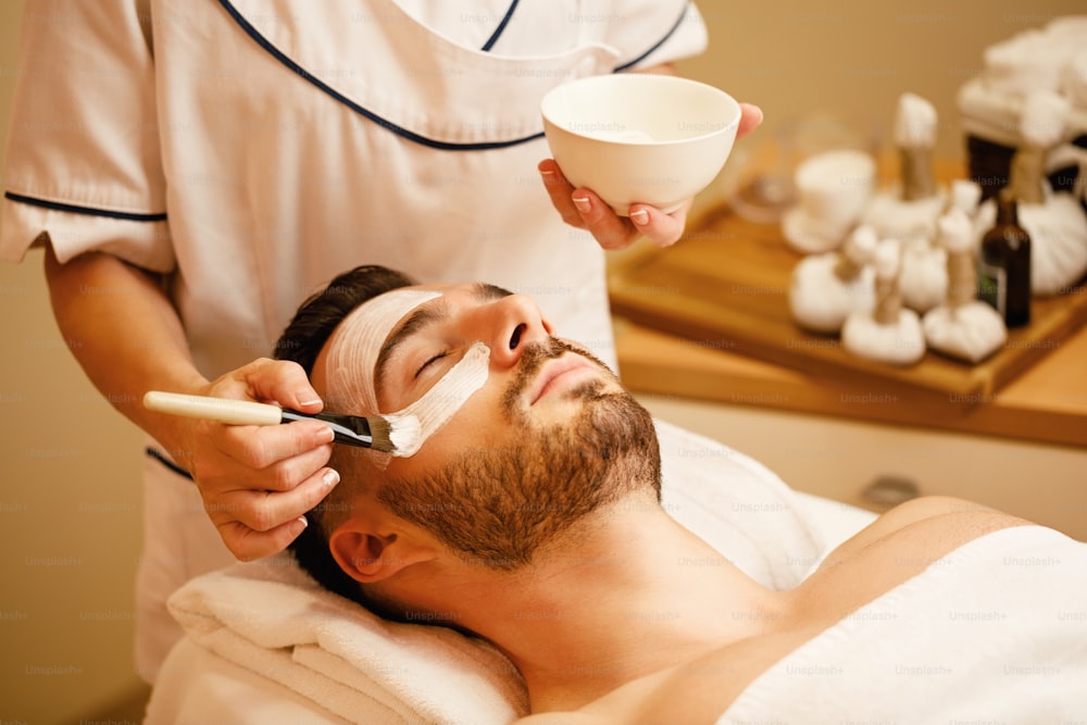 Esteticista irreconhecível aplicando máscara facial no rosto do homem durante o tratamento de spa no centro de bem-estar.